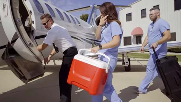 A pilot in a white shirt and a woman in blue scrubs carrying a red cooler walk away from a small airplane while a man pulls a suitcase behind them.