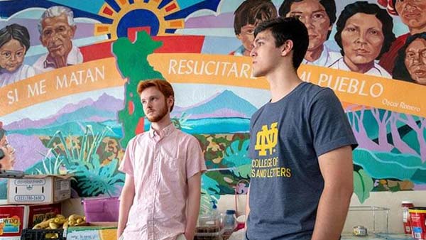 Two young men stand in front of a colorful mural depicting faces, a green landscape, and the phrase 'Si me matan resucitare en el pueblo.' (If they kill me, I will rise in the people.)