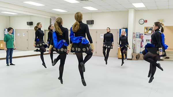Several dancers in black and blue costumes are frozen mid-air as they perform a group dance in a studio with a mirrored wall.