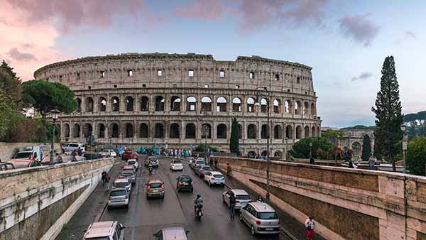 The Colosseum, a large stone amphitheater with arched openings, stands behind a busy street with cars and trees.