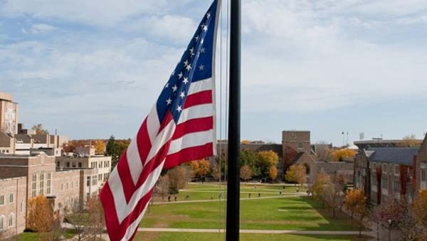 The American flag is at the top of a flagpole on a sunny day overlooking the South Quad on the University of Notre Dame campus.