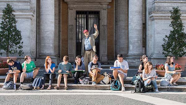 Professor Richard Piccolo gestures with his arm raised while standing behind a group of students sketching on the stone steps of the Square at the top of the Capitoline Hill in Rome, Italy.