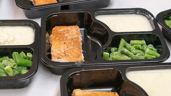 Several black plastic containers hold pre-packaged meals consisting of salmon, green beans, and mashed potatoes.