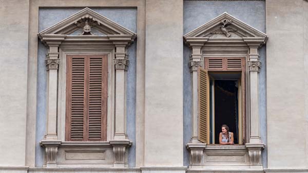 A young woman leans out of an open window with shutters on a stone building with blue and beige walls.