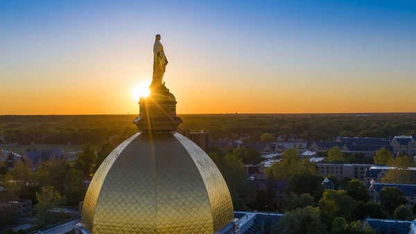 The golden dome and statue of the Virgin Mary on top of the Main Building at the University of Notre Dame are silhouetted against a sunset.
