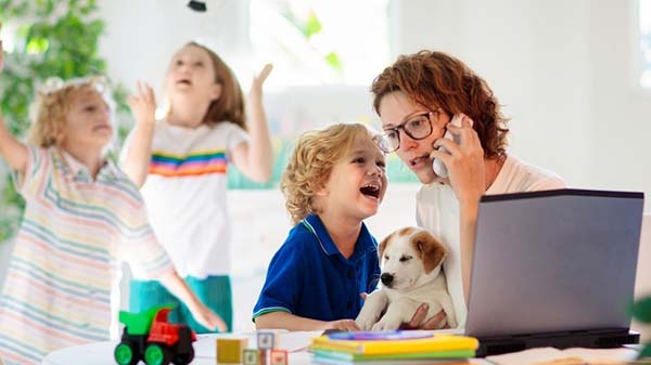 A woman on a phone call works on a laptop at a table with three children and a small dog.