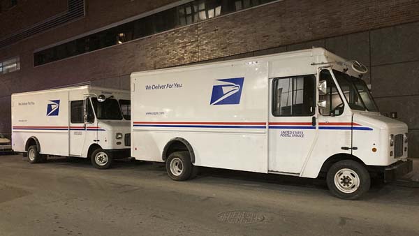 Two United States Postal Service mail trucks are parked side by side on a paved street at dusk.