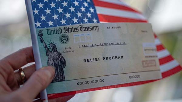 A person's hand holds a United States Treasury stimulus check in front of a small American flag.