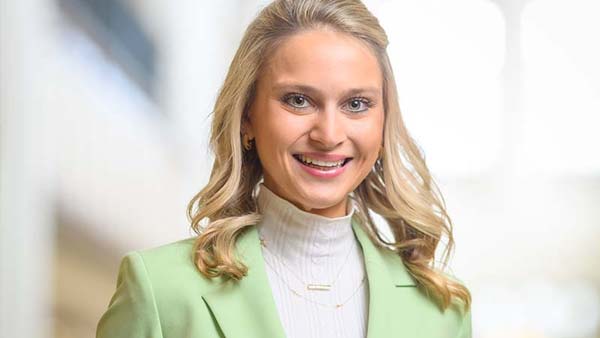 A smiling blonde woman with wavy hair wears a light green blazer and a white turtleneck over a silver necklace.