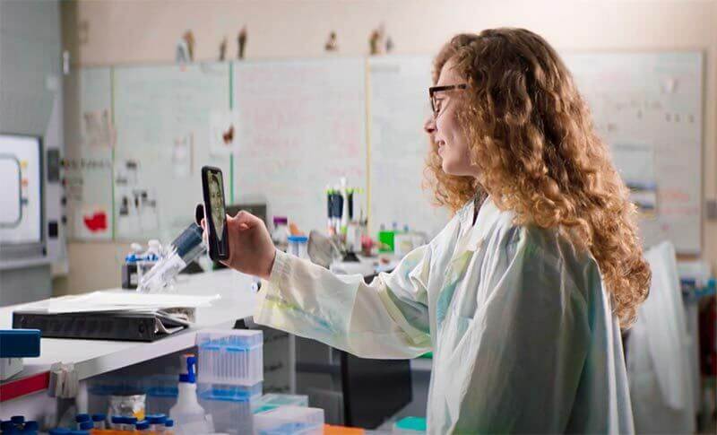 A woman sits in a lab wearing a white lab coat video chatting with another person on her phone.