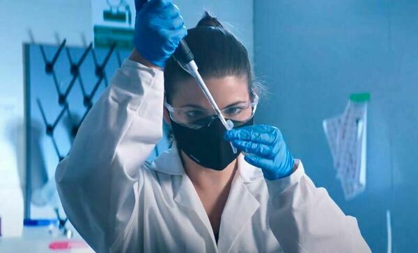A woman wearing a mask, labcoat, gloves, and safety glasses takes a sample from a tube with a syringe.