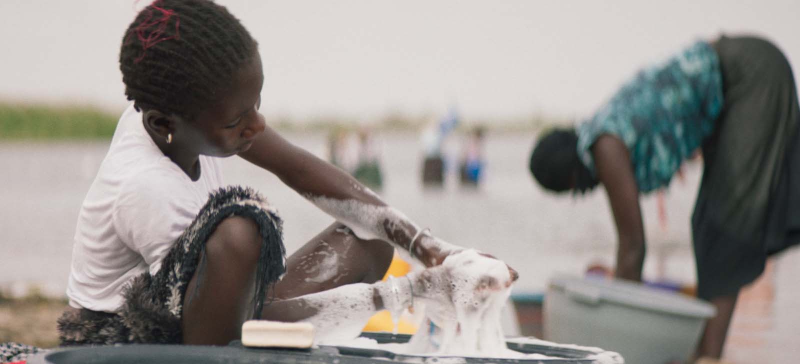 A girl washes clothes in a bucket of soapy water by the river, while a woman does the same in the background. Children swim in the river behind her.