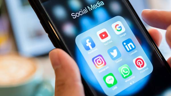 A person's hand holding a smartphone displaying a folder named 'Social Media' containing various app icons, including Facebook, Instagram, YouTube, and Twitter.