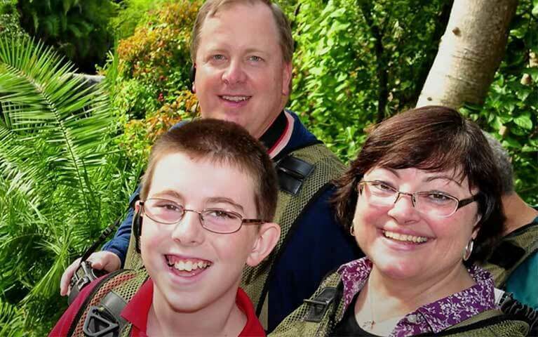 A family photo of three people, a father and mother with their sun smile with tropical trees behind them.