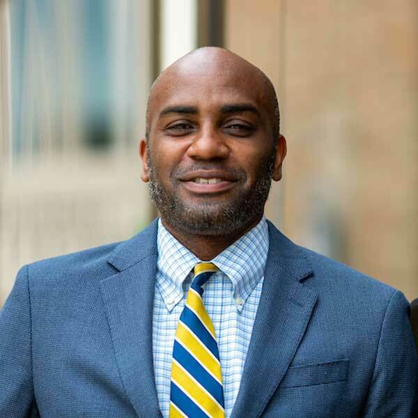 A man standing against a wall wearing a blue suit and blue and yellow stripped tie.