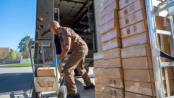 A UPS worker in a brown uniform unloads boxes from the back of a truck onto a hand truck.