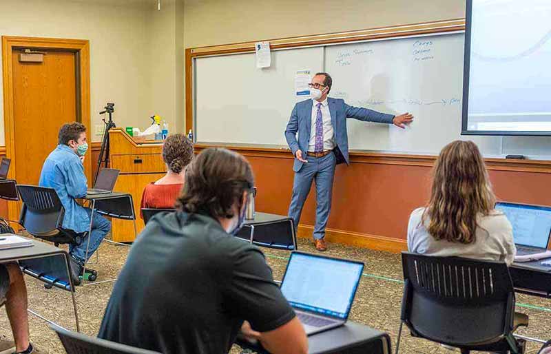 A professor in a suit, tie, and face mask gestures toward a whiteboard while teaching students in a classroom.