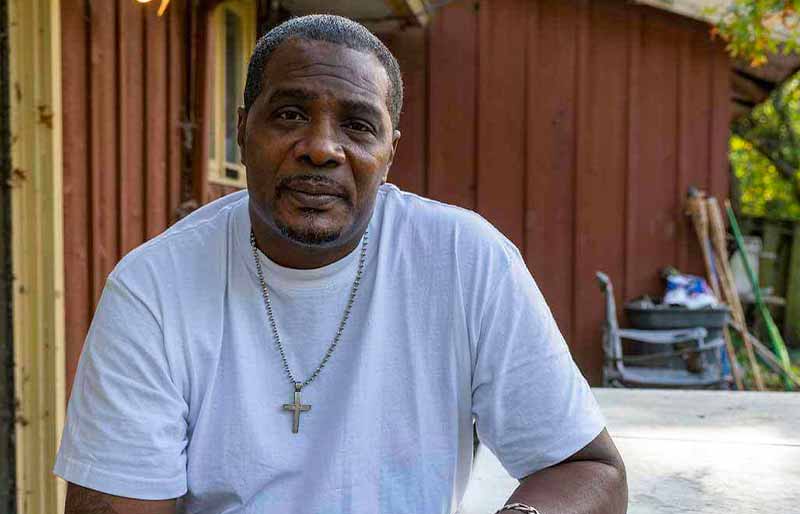 A middle-aged man with a goatee and a silver cross necklace sits outside in front of a house.