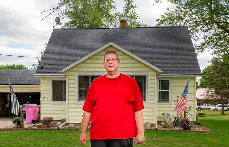 A man with glasses and a red t-shirt stands on a green lawn in front of a small light yellow house with a dark gray roof.
