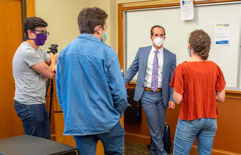 A professor in a suit and tie stands at a whiteboard talking to three students wearing face masks in a classroom.