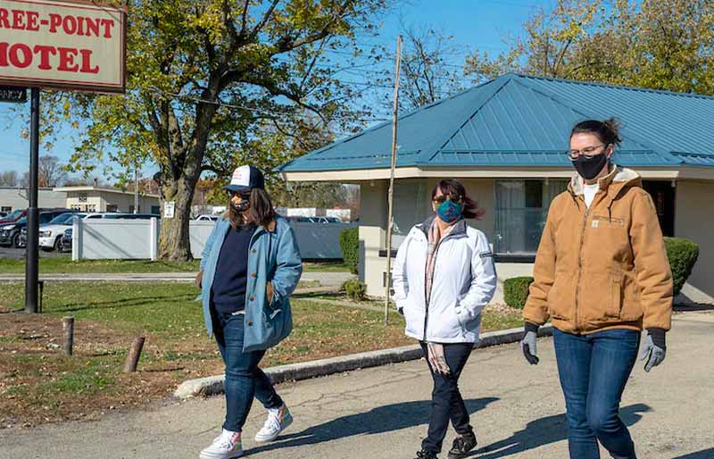 Three women wearing face masks and casual jackets walk along a paved lot in front of the Three-Point Motel.