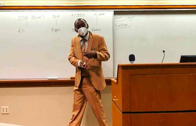 A man in a light brown suit and a white mask stands in front of a whiteboard and a wooden lectern.