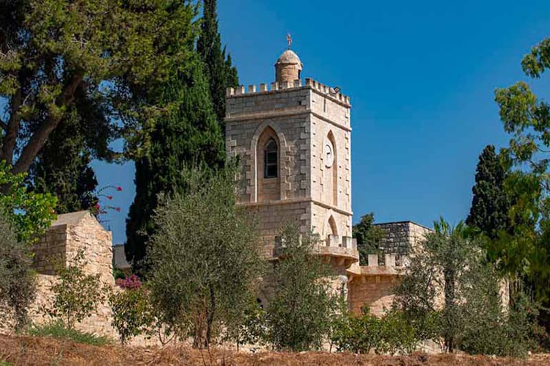 A stone church tower with a clock and cross on top is surrounded by green trees and an old stone wall under a blue sky.
