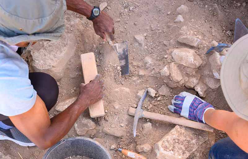 Two archaeologists carefully dig in a rocky, dirt ground using a brush, a small pickax, and a trowel.