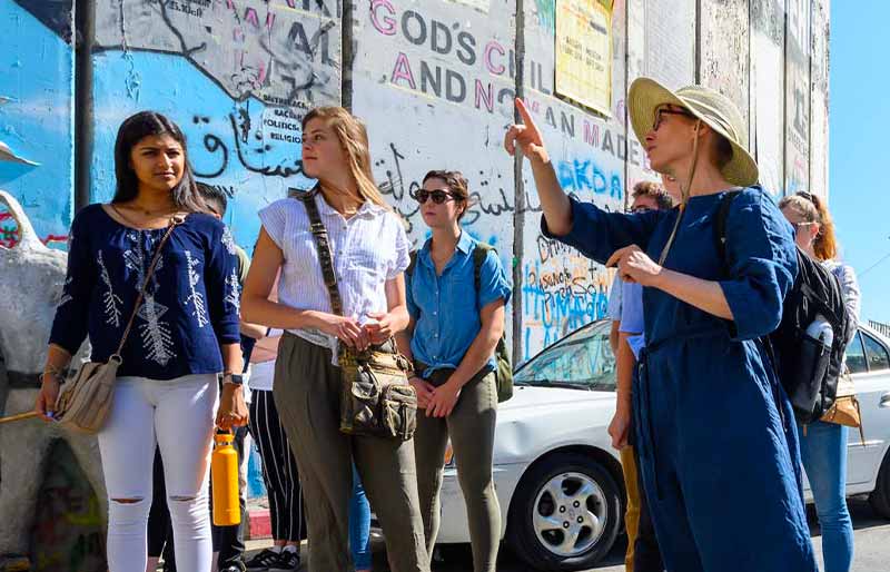 A group of people, including a woman in a sun hat and a blue dress, listen to a tour guide in front of a wall covered in graffiti.
