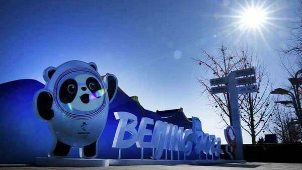 The official Bing Dwen Dwen mascot and a large white 'BEIJING 2022' sign stand outdoors with a bright sun overhead against a clear blue sky.