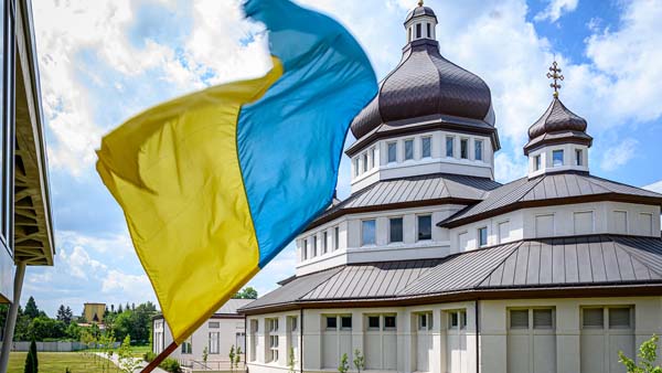 A Ukrainian flag waves in front of a white church with gray domes and crosses on top.