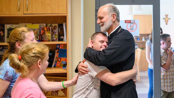 A man with a gray beard and a dark cassock embraces a young man with his eyes closed, as a woman smiles in the background.
