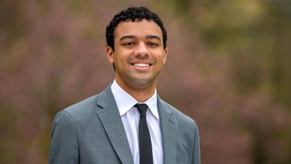 A smiling man with curly dark hair wears a gray suit and a dark blue tie against a soft-focus background of green and pink foliage.