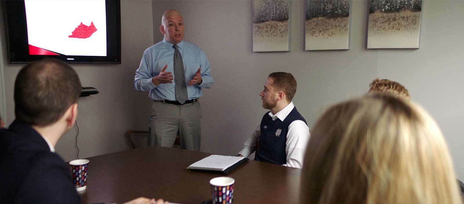 A man stands in front of three people sitting around a table.