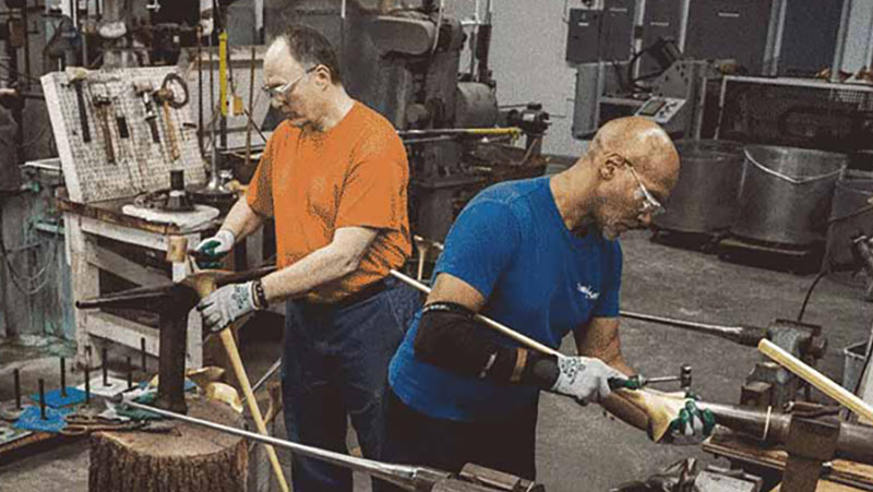 Two men wearing goggles and work gloves work with long metal musical instruments in a workshop.