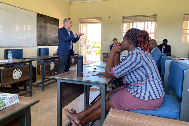 A man in a blue blazer stands and gestures while speaking to a group of students seated at desks in a classroom.
