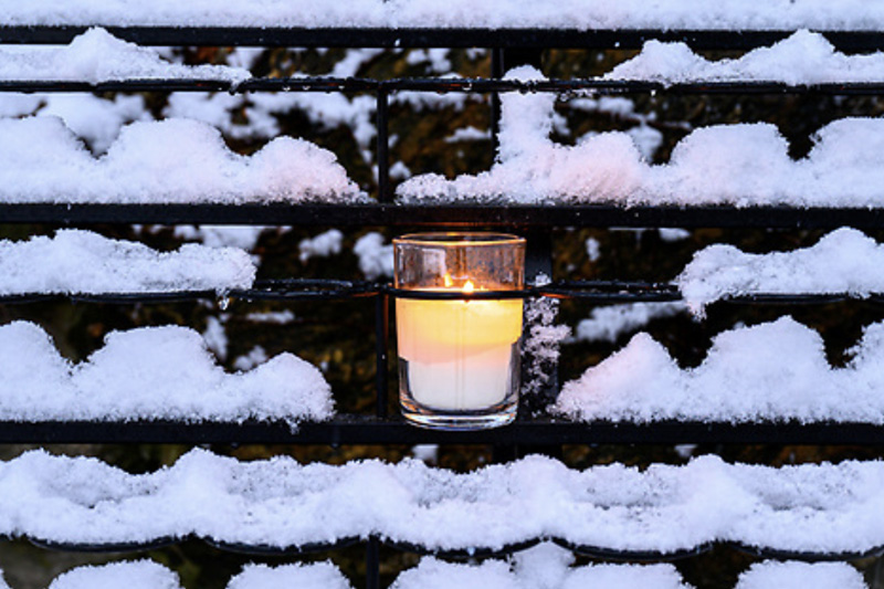 A lit white candle in a glass holder sits on a snowy, black iron grate.