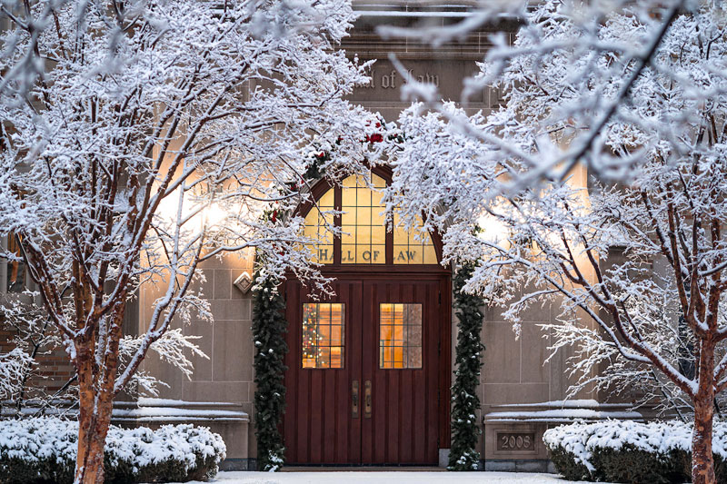 The entrance to Notre Dame Law School on a snowy evening, with the trees and ground covered in fresh white snow and warm light glowing from inside.