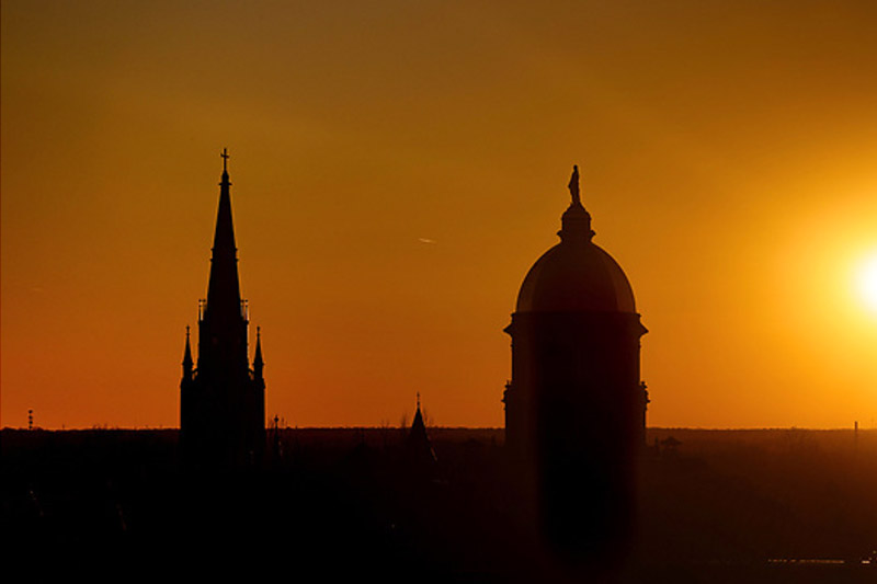 A silhouette of the Sacred Heart Basilica steeple and the Golden Dome against a brilliant golden and orange sunset.