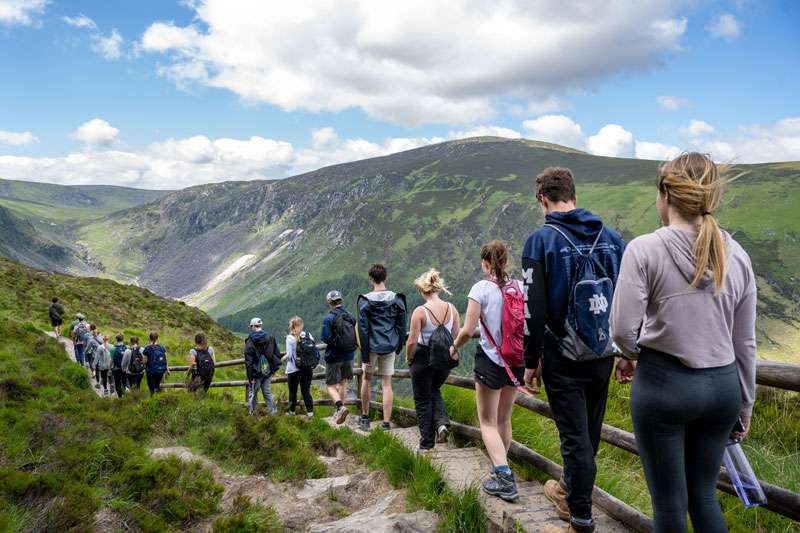 A group of Notre Dame students hike along a path with a wooden railing through a lush, green mountain landscape under a bright, cloudy sky.