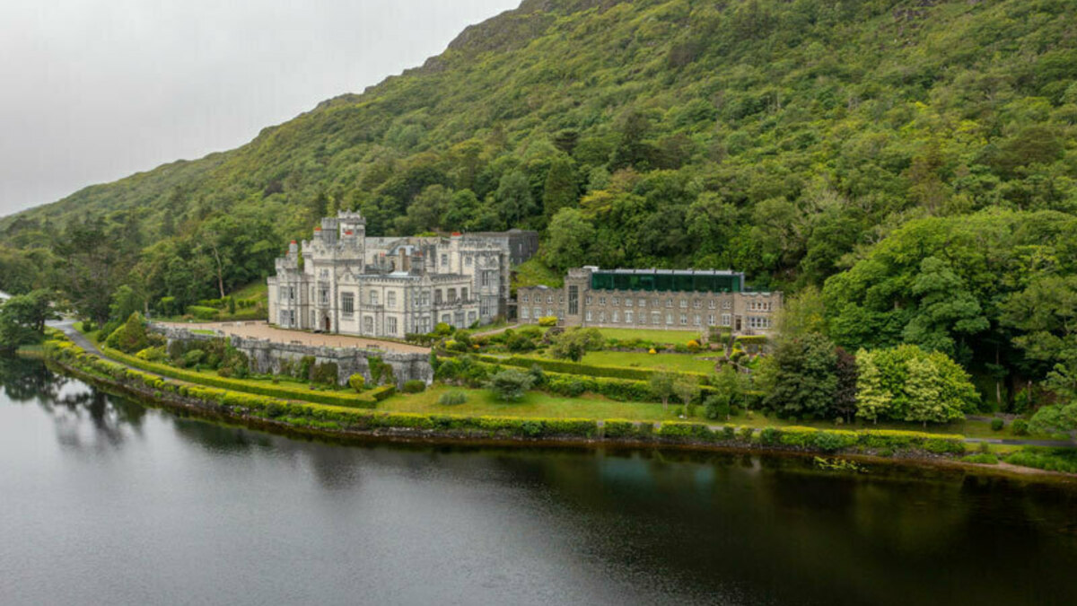 Kylemore Abbey in Ireland sits on a lake shore at the foot of a lush, green, wooded mountain.