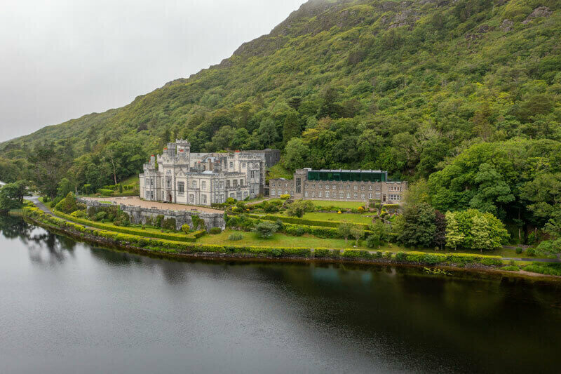 An aerial view of Kylemore Abbey in Ireland, a gray stone building on the edge of a lake, surrounded by green trees and a large, verdant hill.