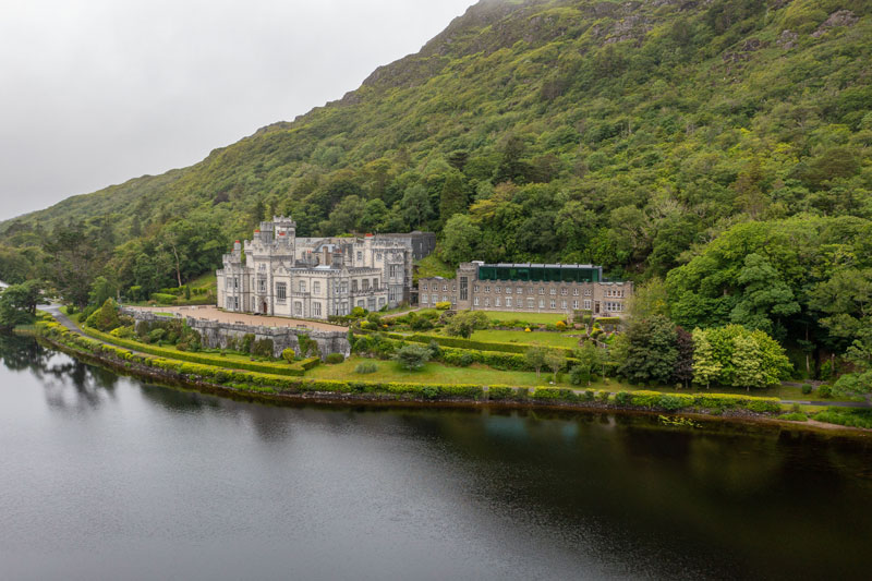 An aerial view of Kylemore Abbey in Ireland, a gray stone building on the edge of a lake, surrounded by green trees and a large, verdant hill.