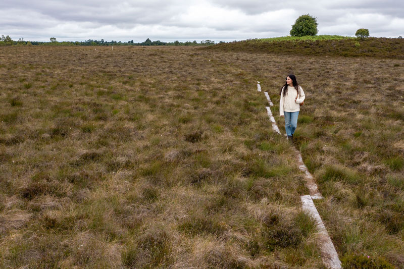 A woman in a white sweater and blue jeans walks along a wooden boardwalk through a field of brown and green grasses under a cloudy sky.