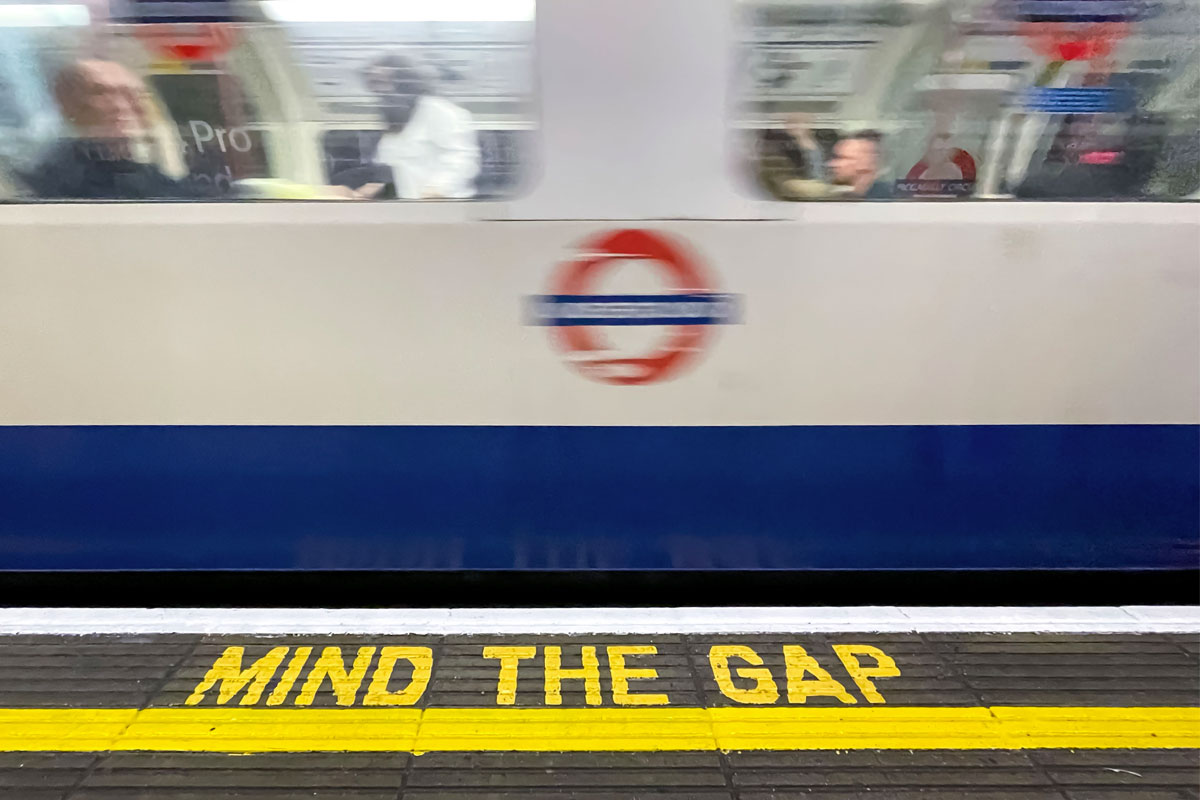A white and blue London Underground train speeds past a platform with the yellow text, 'MIND THE GAP,' painted on the ground.