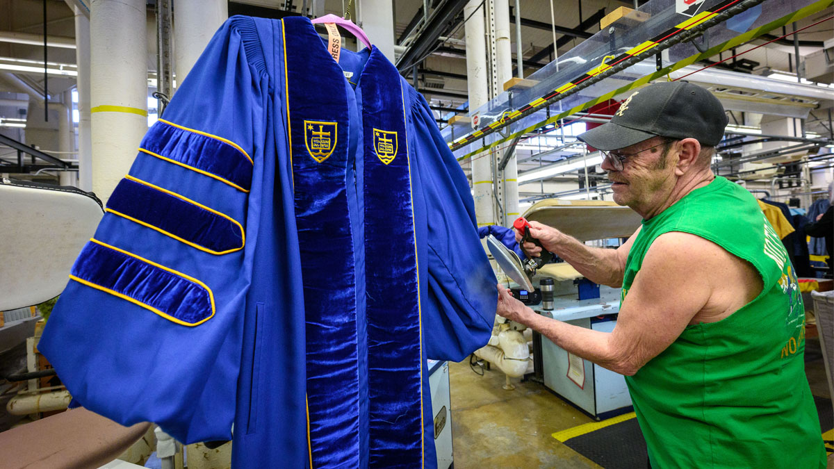 A man in a green shirt and a black baseball cap uses a steaming iron on a Notre Dame doctoral robe in a dry cleaning facility.