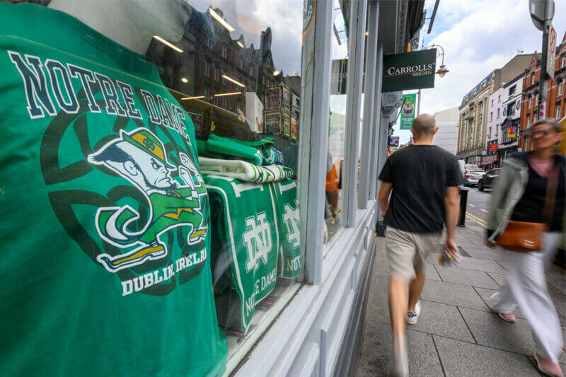 A green Notre Dame Fighting Irish t-shirt is displayed in a store window in Dublin, Ireland, with pedestrians walking by on the street.