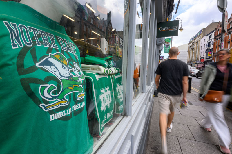 A green Notre Dame Fighting Irish t-shirt is displayed in a store window in Dublin, Ireland, with pedestrians walking by on the street.