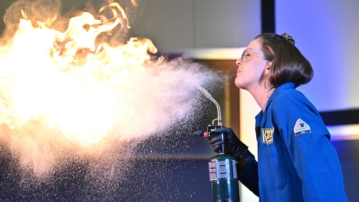 A young female in a blue lab coat and safety goggles stands to the right of the image and faces left. She  holds a gas canister and is seen spitting a flammable liquid into the air. A massive ball of fire fills the left side of the image.