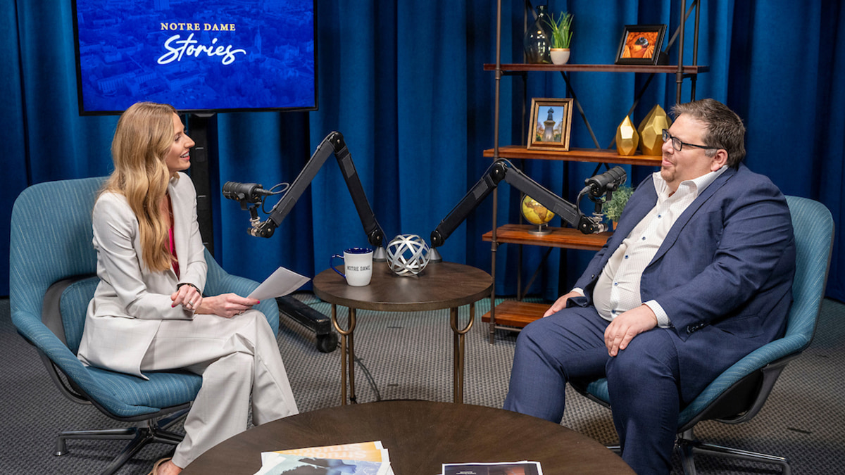 A woman with blonde hair wearing a light gray suit sits next to a man in glasses dressed in a dark blue suit. The two smile at each other. They are separated by a small circular table and two microphones. Behind them is a TV screen that says Notre Dame Stories and a bookcase filled with University-themed decor.